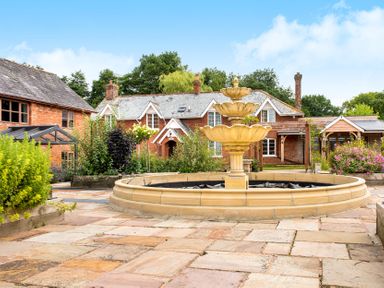 A stone fountain in a paved courtyard with a brick house and garden at Mill Cottage in Aylesbeare