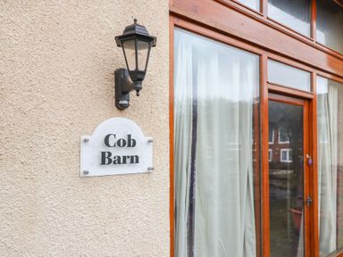 A wall with a light fixture and a sign that says Cob Barn next to a wooden framed glass door with curtains at Cob Barn in Aylesbeare