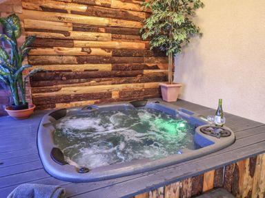 A hot tub with bubbling water beside two potted plants and a bottle of wine with two glasses at Cob Barn in Aylesbeare
