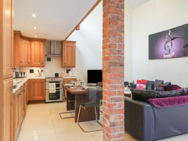 A kitchen with wooden cabinets and a dining table set with cups and plates next to a living area with sofas and a brick pillar at Cob Barn in Aylesbeare