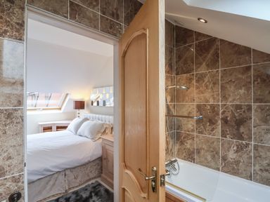 A bedroom with a bed and lamp seen through an open door next to a bathroom with a bathtub and tiled walls at Cob Barn in Aylesbeare