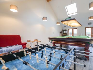 A game room with a foosball table a pool table a red sofa two wooden bar stools and hanging lights at Cob Barn in Aylesbeare