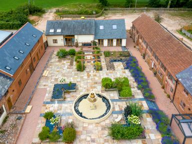 An aerial view of a courtyard with a central circular fountain surrounded by tiled pathways and planted garden beds with buildings on three sides at Cob Barn in Aylesbeare