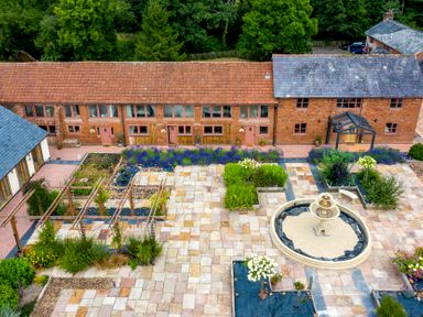 A paved courtyard with a central fountain and various flower beds surrounded by brick buildings and trees at Cob Barn in Aylesbeare