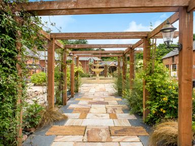 A tiled pathway with wooden pergola and plants leading to a fountain and brick buildings at Cob Barn in Aylesbeare
