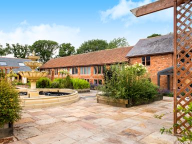 An outdoor courtyard with a tiered stone fountain surrounded by plants and a long brick building with tiled roofs at Cob Barn in Aylesbeare