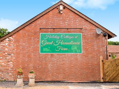 A brick building with a green sign that reads Holiday Cottages at Great Houndbeare Farm and two flower pots on stone stands at Great Houndbeare Farm
