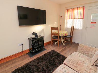 A living room with a wall-mounted TV a black stove a small round table with two wooden chairs a floor lamp and a patterned sofa at Stable Cottage in Cottonshopeburnfoot near Otterburn