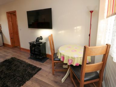 A small dining area with a round table covered with a heart-patterned tablecloth two wooden chairs a black stove a wall-mounted television a floor lamp in a room at Stable Cottage Cottonshopeburnfoot near Otterburn