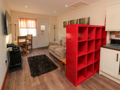 A living area with a patterned sofa a black rug a wooden dining table with chairs a red shelving unit and a wood stove at Stable Cottage in Cottonshopeburnfoot near Otterburn