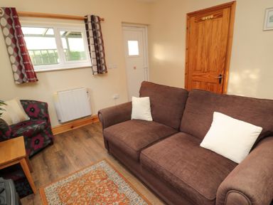A living room with a brown sofa with white cushions a patterned armchair wooden door and window with curtains at Fir Tree Cottage in Cottonshopeburnfoot near Otterburn