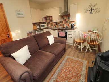 A small living area with a brown sofa and white pillows a kitchen with cabinets a stove and a dining table with two chairs at Fir Tree Cottage in Cottonshopeburnfoot near Otterburn