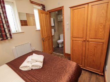 A bedroom with a bed covered with a brown quilt and white towels on it two windows with curtains a wooden wardrobe and an open door leading to a bathroom at Fir Tree Cottage in Cottonshopeburnfoot near Otterburn