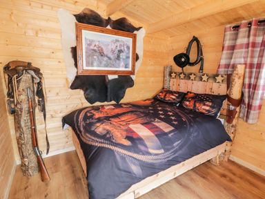 A bedroom with wooden walls and floor a bed with star decorated headboard and cowboy themed bedding a framed painting on a cowhide on the wall a rifle with a saddle and cowboy hat hanging nearby at Whiskey Creek Cabin in Castlerock
