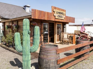A wooden cabin with a sign reading Whiskey Creek a large cactus statue wooden barrels and a wagon with an American flag at Whiskey Creek Cabin in Castlerock