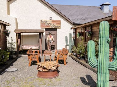 An outdoor western-themed seating area with wooden benches, a fire pit, a swing, and a cowboy statue near a building with a sign that reads Blazin Saddle Saloon at Whiskey Creek Cabin in Castlerock