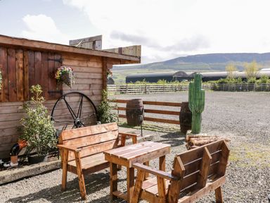 An outdoor seating area with wooden benches and a table next to a wooden building with plants and a wagon wheel