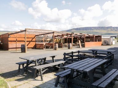 An outdoor seating area with wooden picnic tables and a wooden covered structure with barrels and open sides with mountains in the background at Whiskey Creek Cabin in Castlerock