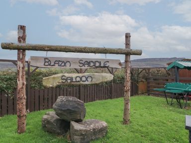 A wooden sign reading Blazin Saddle Saloon hanging between two wooden posts on grass with rocks and a fence in the background at Whiskey Creek Cabin in Castlerock