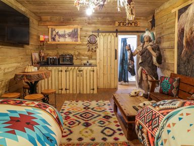 A wooden room with beds, a colorful rug, a wooden table with stools, a kitchenette, and a large statue of a native american at Apache Falls Lodge in Castlerock