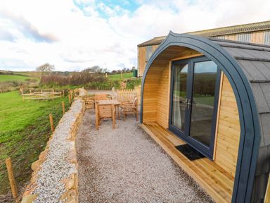 A wooden pod with glass double doors and outdoor wooden furniture on gravel near a stone wall at Audrey in Penzance near Newlyn