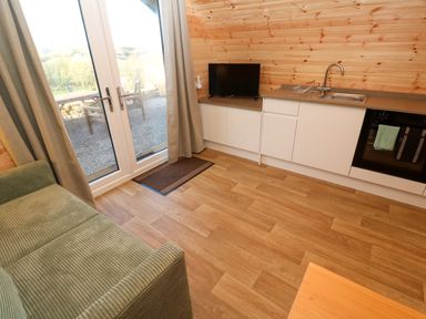 A kitchen area with white cabinets a sink a small television a green sofa and glass doors leading outside at Audrey in Penzance near Newlyn