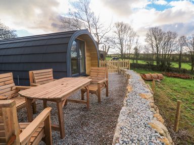 An outdoor seating area with wooden table and chairs next to a curved cabin and stone wall in a rural setting at Audrey in Penzance near Newlyn