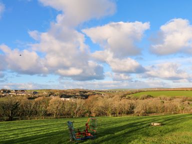A green field with a piece of farming equipment and a pile of stones with trees and houses in the background under a blue sky with clouds at Audrey in Penzance near Newlyn
