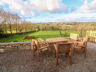 An outdoor seating area with wooden table and chairs overlooking a green field with trees and farm equipment at Lloyd in Penzance near Newlyn