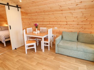 A small dining area with a wooden table and four chairs next to a green sofa and a bedroom with a bed behind a sliding door at Lloyd in Penzance near Newlyn