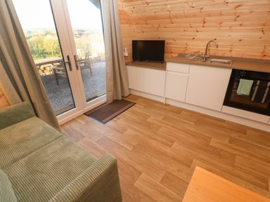 A small kitchen area with white cabinets a sink a black oven and a television next to glass doors with curtains and a green sofa at Lloyd in Penzance near Newlyn