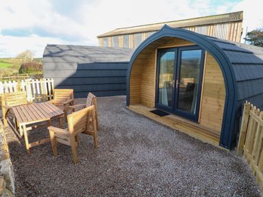 An outdoor seating area with wooden table and chairs on gravel in front of a curved wooden cabin with glass doors at Lloyd in Penzance near Newlyn