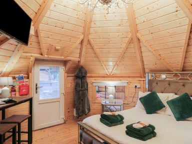 A wooden bedroom with a bed, green pillows and towels, a bedside table with a lamp, a door, a wall-mounted TV, and a hanging robe at Hexagon Hut - Dec Update in Dungiven Londonderry
