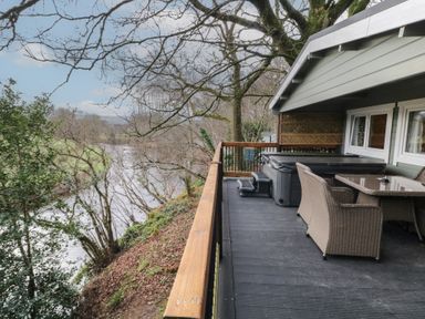 An outdoor deck with wicker chairs, a glass table and a hot tub overlooking a river and trees at Mill View in Dungiven Londonderry