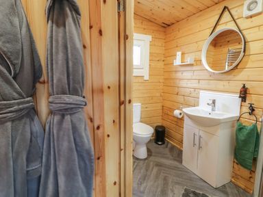 A bathroom with wooden walls a toilet a white sink with cabinet a round mirror a green towel and two gray bathrobes at Owenbeg Dungiven Londonderry