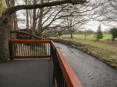 A wooden deck with railing beside a flowing river and trees at Juniper in Dungiven Londonderry