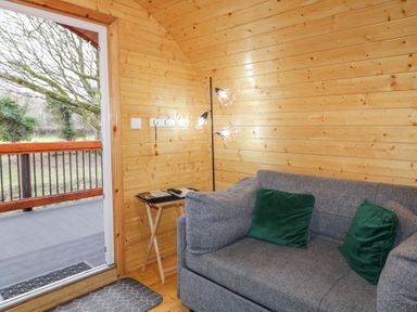 A room with wooden walls featuring a grey sofa with green cushions a small side table with reading materials and a floor lamp next to an open door leading to an outdoor area at Yew in Dungiven Londonderry