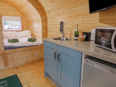 A wooden cabin interior with a bed with white bedding and green towels next to a blue kitchen cabinet with a sink microwave and mini fridge at Yew in Dungiven Londonderry