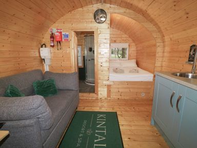 A wooden cabin interior with a gray sofa green cushions kitchenette bed with towels and a shower at Kintail Resort Spa