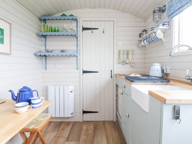 A small kitchen with wooden floors wooden shelves with dishes and glasses a wooden table with a blue teapot and mugs a door in the center and a kitchen sink on the right at Teg in Prospidnick near Porthleven