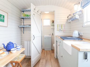 A kitchen with a wooden table and stools a blue teapot cups shelves with glassware and plates a door leading to a small bathroom at Teg in Prospidnick near Porthleven