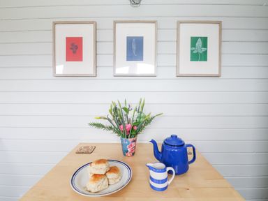 A wooden table with a plate of scones a blue teapot a creamer jug and a floral vase in a room with three framed botanical prints on the wall at Teg Prospidnick near Porthleven