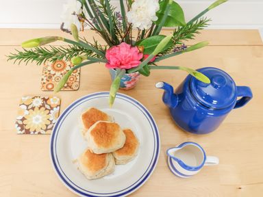 A plate with four biscuits next to a blue teapot and small creamer on a wooden table with flower coasters and a floral arrangement at Teg in Prospidnick near Porthleven