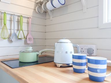 A kitchen counter with a green pot on a stove a white SMEG kettle two blue and white mugs and cooking utensils hanging on a white wall at Teg in Prospidnick near Porthleven