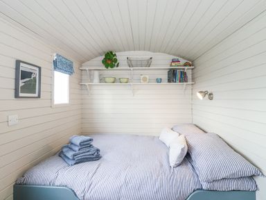 A small bedroom with a bed with striped bedding folded towels on the bed shelves with books plants and dishes on the wall at Teg in Prospidnick near Porthleven