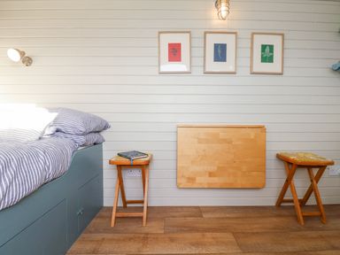 A bedroom with a bed with striped bedding two wooden stools one with a book a wall-mounted wooden foldable table and three framed pictures at Teg in Prospidnick near Porthleven