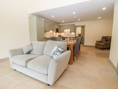 An open plan living and dining area with a gray sofa and armchair next to a dining table and kitchen at Bears Court Barn 3 Little Rissington near Bourton-On-The-Water