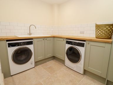 A laundry room with washing machine, dryer, sink, green cabinets, wooden countertop, and a laundry basket at Bears Court Barn 3 Little Rissington near Bourton-On-The-Water