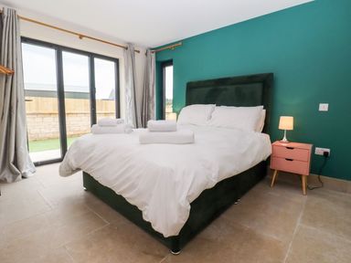 A bedroom with a double bed, white bedding, towels on the bed, a side table with a lamp, teal accent wall, and sliding glass doors with gray curtains at Bears Court Barn 3 Little Rissington near Bourton-On-The-Water