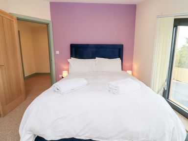 A bedroom with a bed with white linens and two rolled towels bedside lamps and a purple accent wall Bears Court Barn 3 Little Rissington near Bourton-On-The-Water